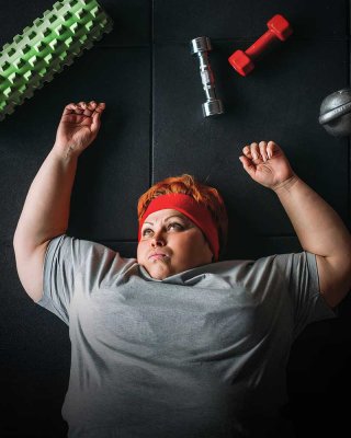 Женщина, лежащая на полу в спортзале в окружении спортивного инвентаря, выглядит задумчивой и разочарованной. Это визуализирует распространенное чувство, когда усилия по подсчету калорий не приносят желаемого результата, о чем и пойдет речь в статье.