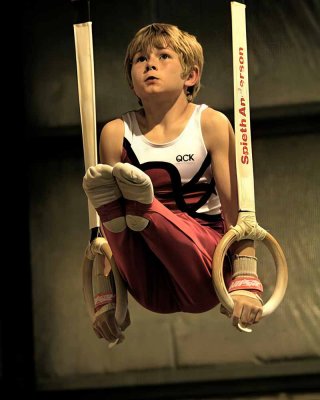 Ребёнок выполняет упражнение на гимнастических кольцах, напряжённые мышцы и внимательный взгляд передают атмосферу первых серьёзных тренировок и становления маленького спортсмена. Фото дополняет материал о том, когда лучше начинать занятия спортивной гимнастикой для мальчиков и как спорт развивает ловкость, силу и уверенность.